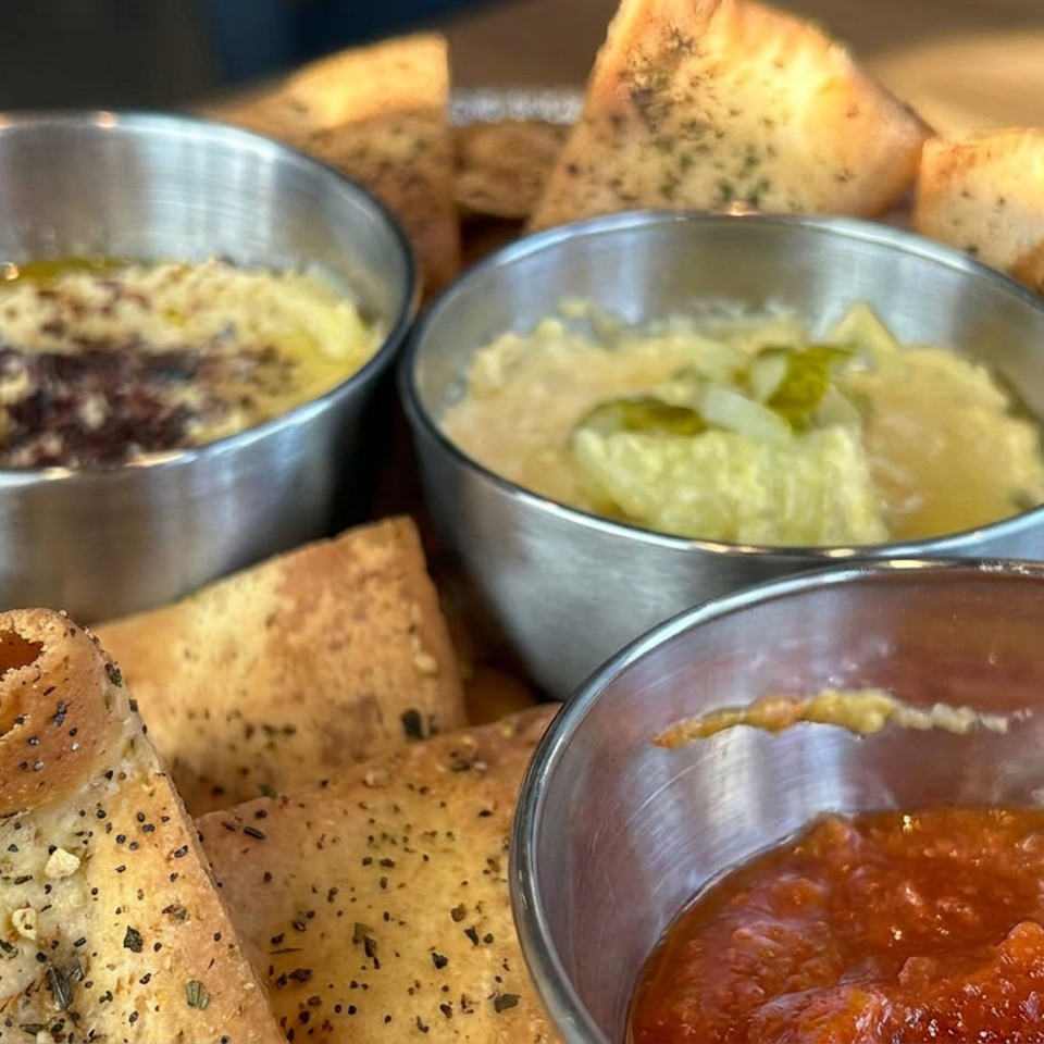 Assorted seasoned flatbread chips surround three metal bowls of creamy dips, guacamole, and red salsa on a wooden table.