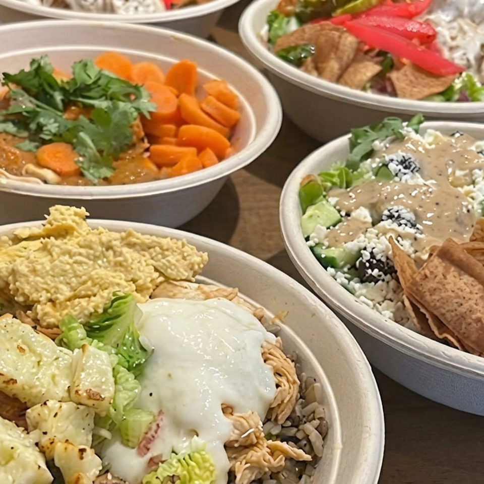 Several bowls of Mediterranean-inspired meals feature hummus, grilled chicken, rice, fresh vegetables, pita, sauces, and colorful salads on a wooden table.